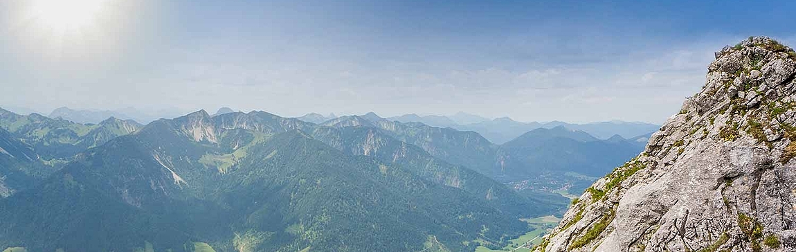 Eine weite Panoramaaufnahme einer bergigen Landschaft. Im Vordergrund rechts ragt ein felsiger Gipfel empor. Links davon erstrecken sich grüne Täler und bewaldete Berge unter einem strahlend blauen Himmel mit der Sonne.